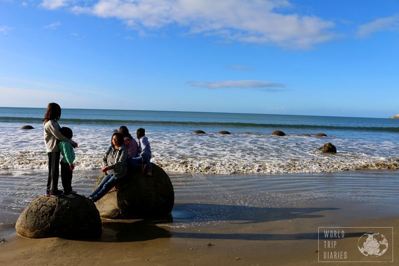 2 round boulders half sunk in the sand of the beach at Moeraki beach, in NZ, and 2 kids standing on one and 3 people sitting on the other. Moeraki is a wonder!